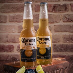 Cold Corona Extra beer bottles on a wooden table with lime slices against a brick wall backdrop.